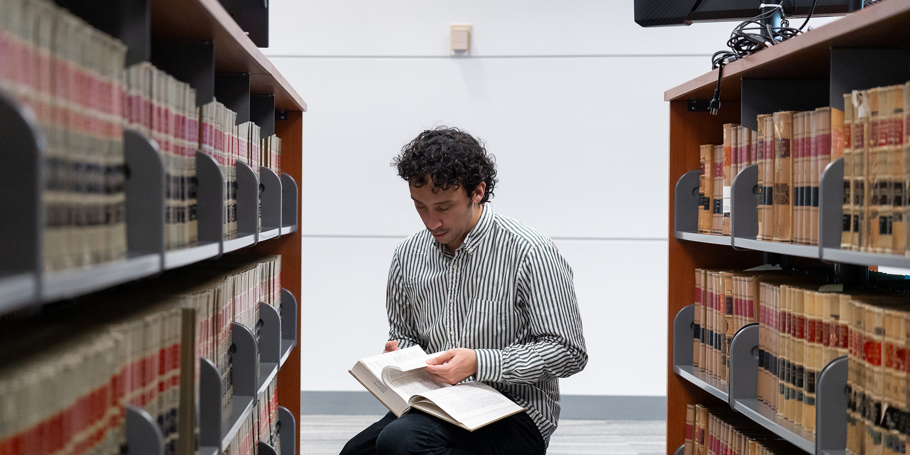 student sitting between book shelves, looking down at an open book