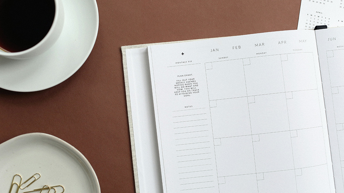 Cup of coffee on desk in top left corner, dish with paper clips at bottom left corner, with date organizer open at a diagonal view